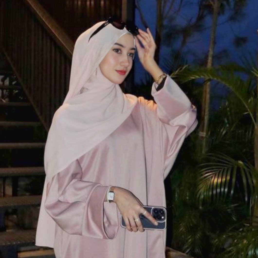 Woman in a light pink outfit with a hijab taking a selfie outdoors.